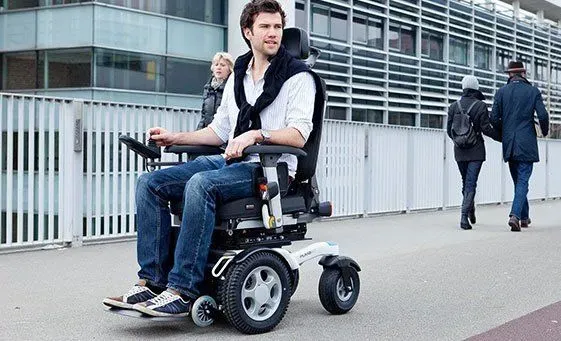 A man in jeans with a sweater around his shoulders steering an electric wheelchair down a downtown sidewalk